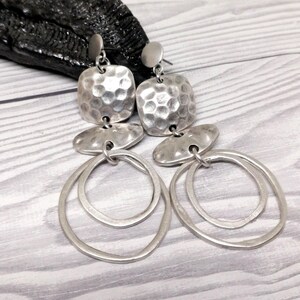 Silver earrings featuring a hammered square top, an oval middle section, and two nested oval hoops. The earrings have a post backing and an antique finish. The jewellery is displayed on a light grey wooden surface.