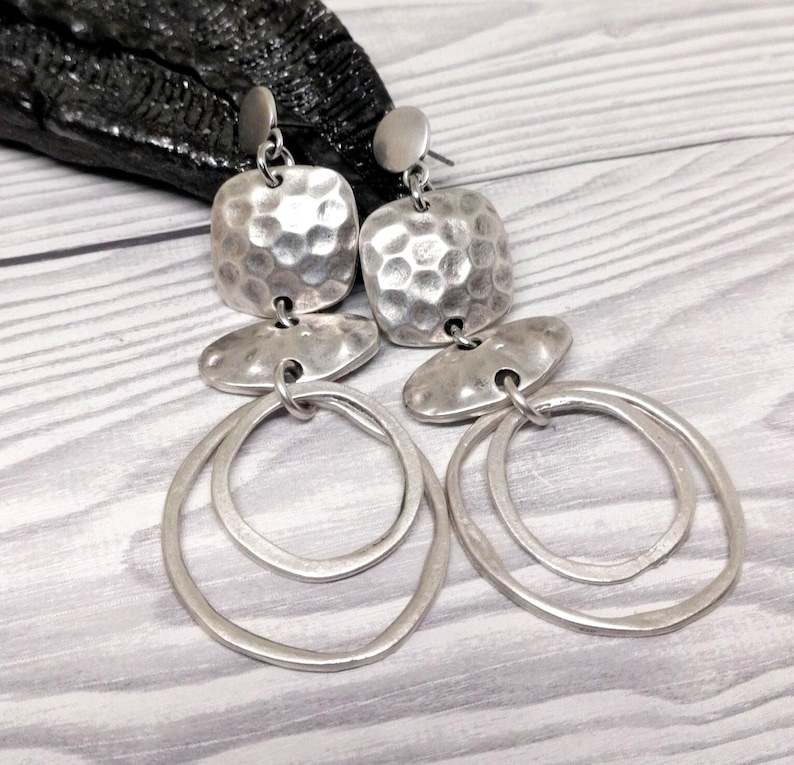 Silver earrings featuring a hammered square top, an oval middle section, and two nested oval hoops. The earrings have a post backing and an antique finish. The jewellery is displayed on a light grey wooden surface.