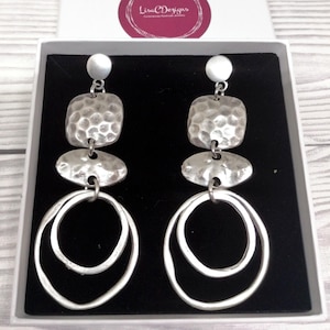 Silver-coloured statement earrings with a layered design. Each earring features a small round stud, a textured square, an oval shape, and two connected open circles. The earrings are displayed in a white box with a black interior.