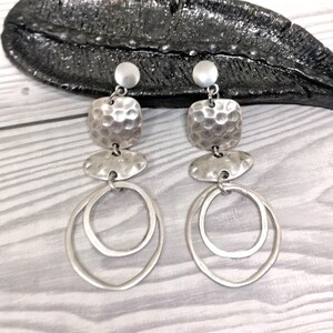 A pair of silver earrings. Each earring has a small round stud, a textured square, a flat oval, and two overlapping oval hoops. The earrings are presented on a light wood-grain surface.