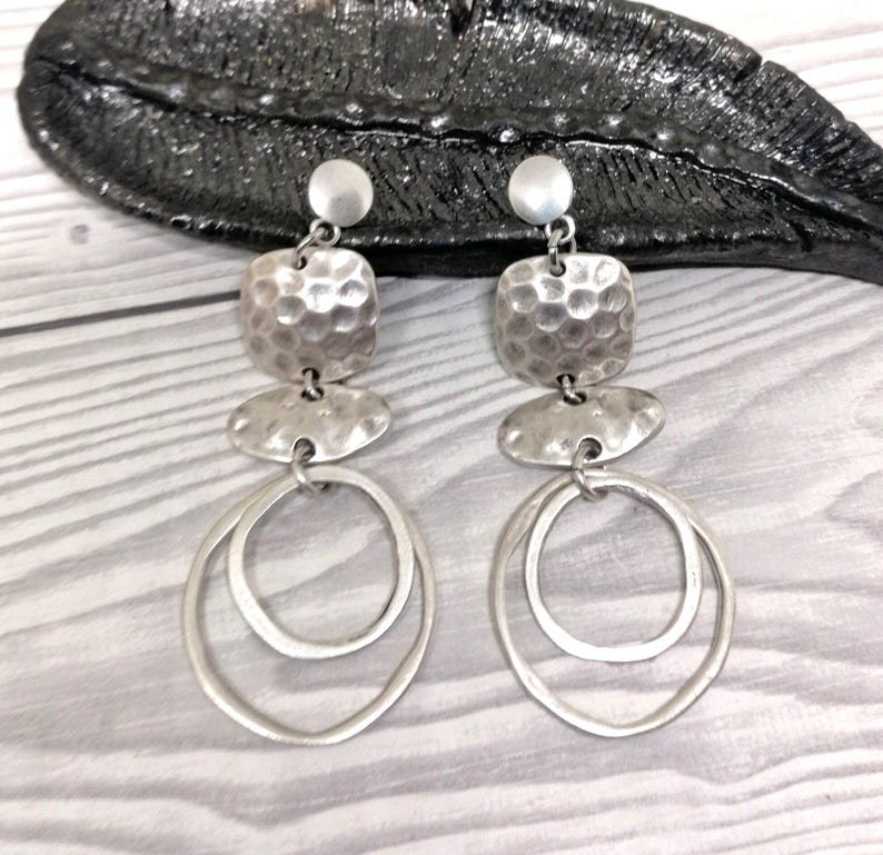 A pair of silver earrings. Each earring has a small round stud, a textured square, a flat oval, and two overlapping oval hoops. The earrings are presented on a light wood-grain surface.