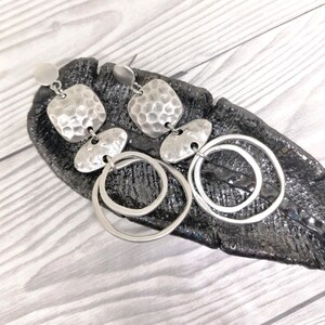 Silver-coloured drop earrings with a hammered texture and circular details. The earrings have a post back and are presented on a dark, textured surface. The design incorporates a mix of geometric shapes.