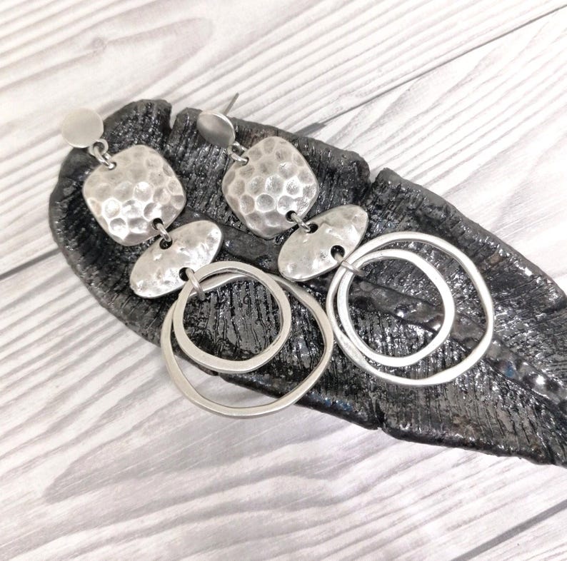 Silver-coloured drop earrings with a hammered texture and circular details. The earrings have a post back and are presented on a dark, textured surface. The design incorporates a mix of geometric shapes.