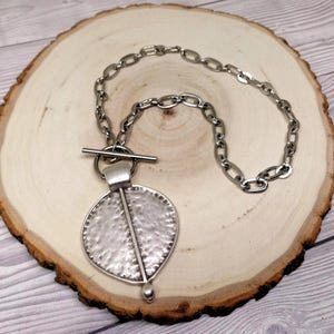 May include: A silver-toned necklace featuring a large, circular pendant with a hammered, textured surface and a vertical bar detail. The pendant is attached to a thick, oval-link chain that secures with a horizontal bar and ring toggle clasp.
