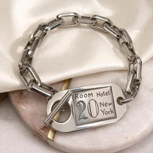 May include: A silver chain bracelet featuring a rectangular tag with the text "ROOM Hotel 20 New York". The bracelet is made of linked chain pieces and has a toggle clasp. The tag is in a classic font.