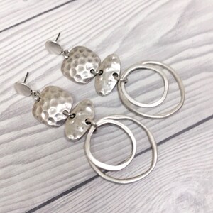 A pair of silver earrings. Each earring has a textured, hammered-metal design with two irregular shapes and two connected circular hoops. The earrings are displayed on a light-coloured wooden surface.