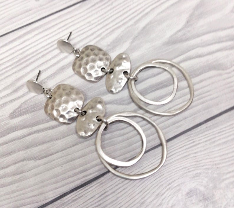 A pair of silver earrings. Each earring has a textured, hammered-metal design with two irregular shapes and two connected circular hoops. The earrings are displayed on a light-coloured wooden surface.