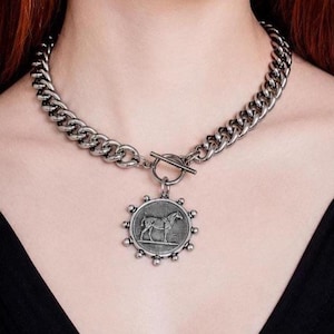 May include: Silver-coloured chain necklace with a circular pendant depicting a horse. The necklace features a thick chain design and a toggle clasp. The pendant is detailed with small, round embellishments around the edge.