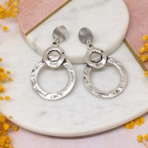 May include: A pair of silver-toned drop earrings featuring a textured, hammered metal finish. Each earring consists of a small circular stud connected to a larger open ring pendant, creating a layered, dangling design with a matte, metallic appearance.