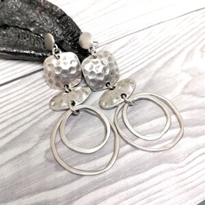 Silver-coloured dangle earrings. Each earring features a hammered square top, an oval middle piece, and two overlapping circular hoops. The earrings have a textured, metallic finish. Displayed on a light grey wooden surface.