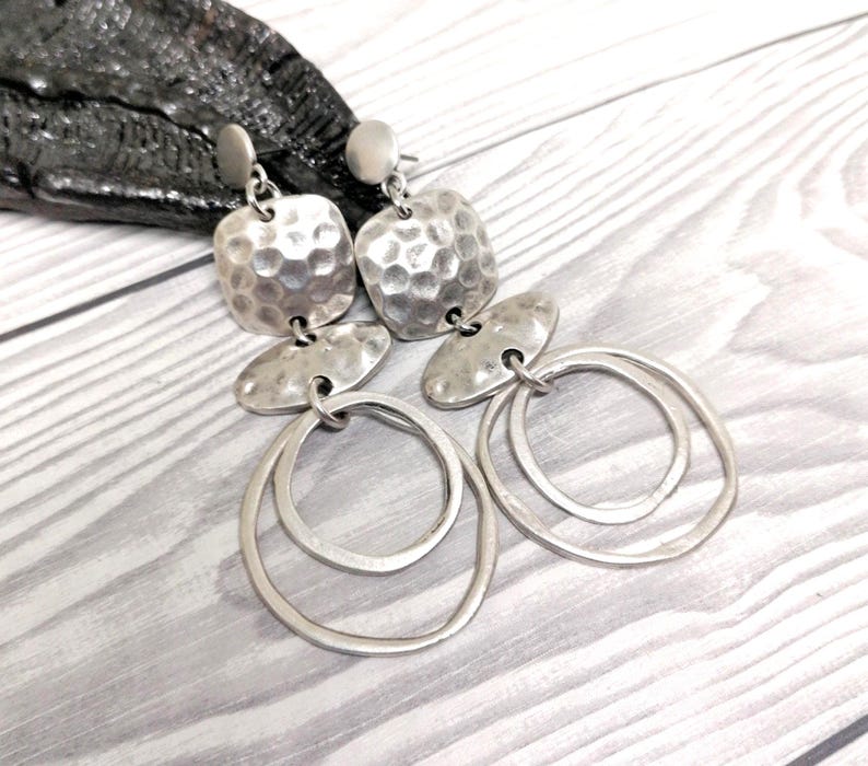 Silver-coloured dangle earrings. Each earring features a hammered square top, an oval middle piece, and two overlapping circular hoops. The earrings have a textured, metallic finish. Displayed on a light grey wooden surface.