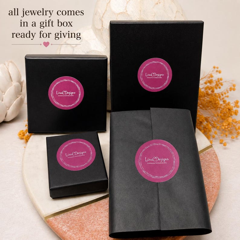 Four black gift boxes and a wrapped package, each with a pink circular sticker that reads "Lisa Designs". The text "all jewelry comes in a gift box ready for giving" is at the top of the image.