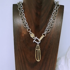 May include: A double-strand silver-toned chain necklace featuring a textured finish. The necklace includes a gold-toned circular charm with a cross design, a silver-toned U-shaped shackle clasp, and a long, vertical gold-toned oval pendant.