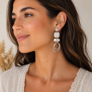 Silver-coloured dangle earrings with a geometric design. Each earring features a small round stud, a textured square, an oval shape, and two connected oval rings. The earrings are worn on a person.
