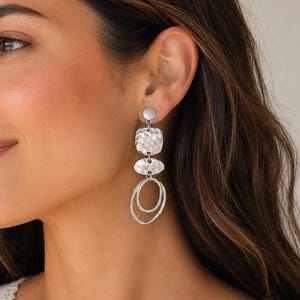 Silver drop earrings with a hammered texture and a layered design. The earrings feature a small circle at the top, a square shape, an oval shape, and two connected oval shapes at the bottom. The earrings are a modern accessory.