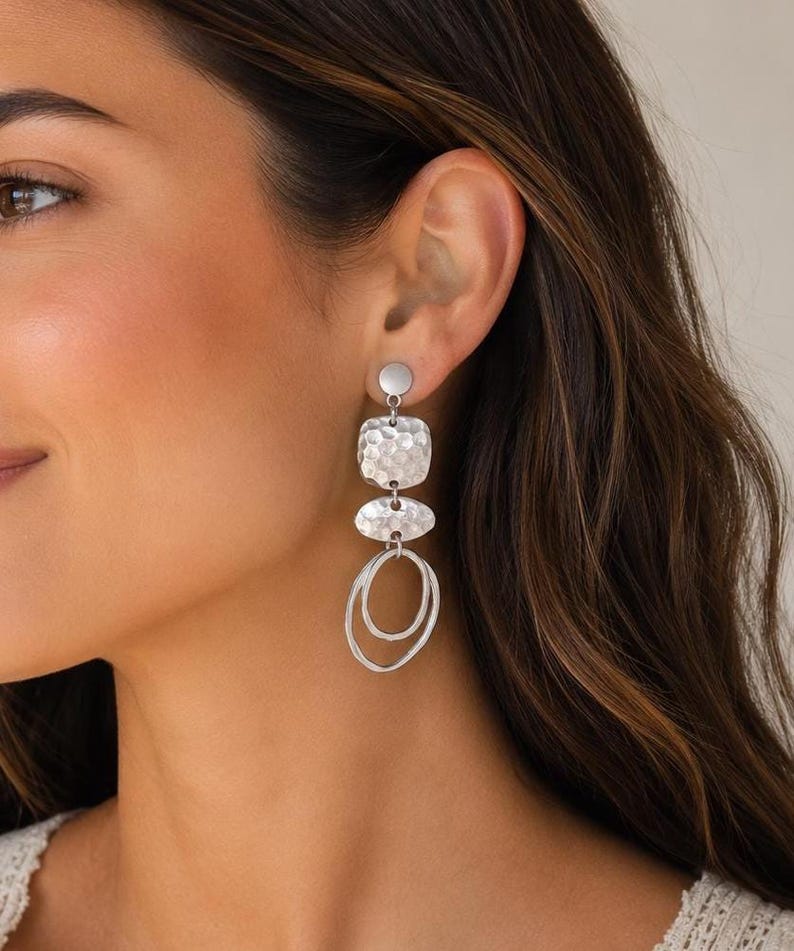 Silver drop earrings with a hammered texture and a layered design. The earrings feature a small circle at the top, a square shape, an oval shape, and two connected oval shapes at the bottom. The earrings are a modern accessory.