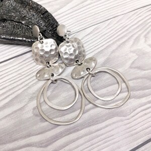 Silver-coloured drop earrings featuring a hammered square top, an oval middle section, and two overlapping circular hoops. The earrings are presented on a light wood-grain surface.