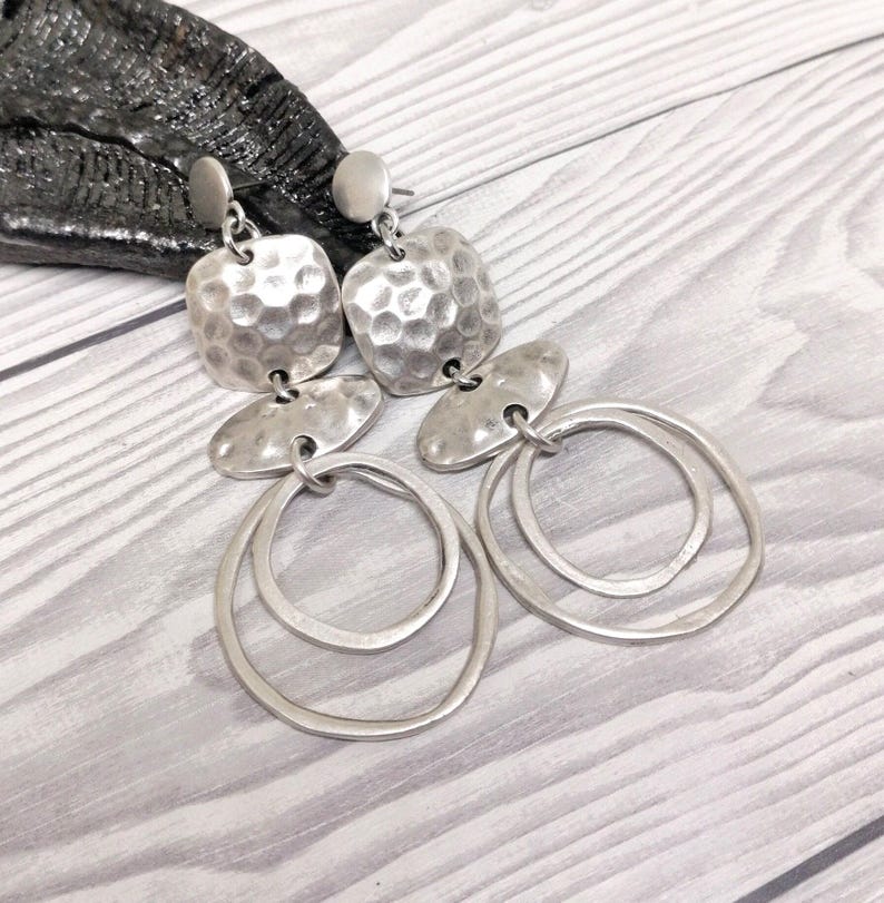 Silver-coloured drop earrings featuring a hammered square top, an oval middle section, and two overlapping circular hoops. The earrings are presented on a light wood-grain surface.