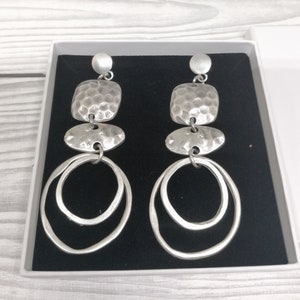 Silver-coloured statement earrings with a layered design. Each earring features a small round stud, a hammered square, an oval shape, and two large, overlapping hoops. The earrings are presented in a white box with a black interior.