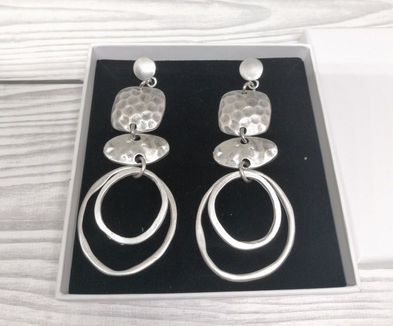 Silver-coloured statement earrings with a layered design. Each earring features a small round stud, a hammered square, an oval shape, and two large, overlapping hoops. The earrings are presented in a white box with a black interior.