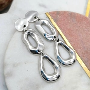 May include: Silver-coloured drop earrings with a contemporary, abstract design. Each earring has a small, round stud connected to three open, organic shapes. The earrings have a polished finish and hang from a small metal loop.