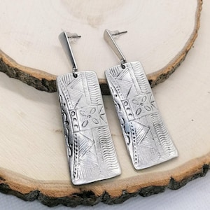 May include: A pair of silver-coloured rectangular earrings with a post back. The earrings have an embossed geometric pattern with floral and linear designs. The earrings are displayed on a wooden surface.