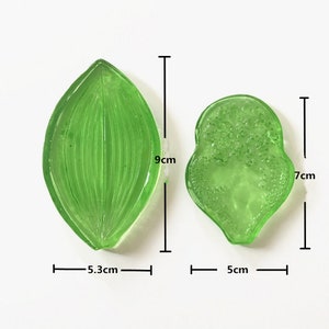 Könnte beinhalten: Zwei grüne Kunststoff-Blätterformen zum Backen oder Basteln. Das größere Blatt ist 9 cm lang und 5,3 cm breit. Das kleinere Blatt ist 7 cm lang und 5 cm breit.