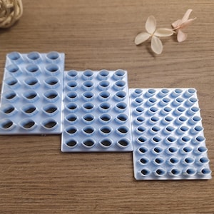 Può includere: Tre strumenti artigianali in plastica blu chiaro con vari ritagli a forma ovale. Gli strumenti sono disposti su una superficie di legno, con un fiore secco e una palla intrecciata sullo sfondo. Questi strumenti sono probabilmente utilizzati per l'artigianato o la decorazione.