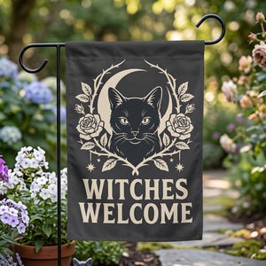 Könnte beinhalten: Eine rechteckige Gartenflagge mit schwarzem Hintergrund und einer cremefarbenen Grafik einer Katze, die von einer Mondsichel und einem Rosenkranz umgeben ist. Der Text "WITCHES WELCOME" steht in fetten, cremefarbenen Großbuchstaben unter der Katze.