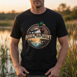 May include: A black t-shirt featuring a circular graphic with a mallard duck at the top and a hunter in a blind by a lake at the bottom. The text BORN TO HUNT is printed in a light tan font across the centre of the circular wooden-textured frame.