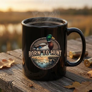 Peut inclure: Un mug en c&eacute;ramique noire rempli d'une boisson sombre, orn&eacute; d'un motif circulaire repr&eacute;sentant un canard colvert, un chasseur dans un aff&ucirc;t et le texte BORN TO HUNT. Le design inclut un coucher de soleil sur un lac avec des roseaux.