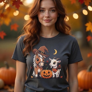 May include: Dark gray t-shirt featuring a Halloween-themed graphic. The design includes a cartoon horse wearing a witch hat, a goat with a bandana, a jack-o'-lantern, and a crescent moon. The background shows pumpkins and autumn foliage.