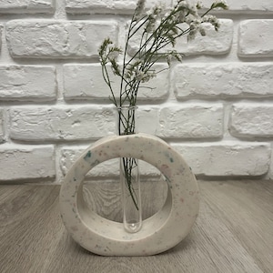 Può includere: Vaso in ceramica bianca con un'apertura rotonda e un tubo di vetro trasparente all'interno. Il vaso ha un motivo maculato ed è pieno di fiori bianchi.