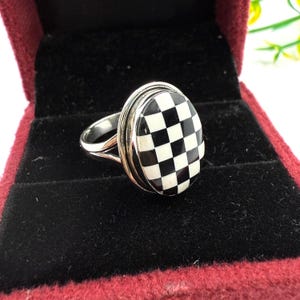 May include: A silver ring with an oval-shaped, black and white checkered design. The ring is set in a red velvet jewelry box.