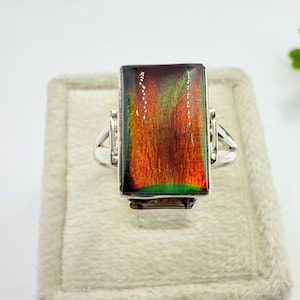 May include: A rectangular gemstone ring with a silver band. The gemstone displays a vibrant array of colors, including red, orange, and green. The ring is set on a light beige surface, showcasing the jewelry's design and color.