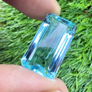 May include: A rectangular, emerald-cut gemstone with a transparent, light blue color. The stone features faceted edges that reflect light, creating a clear, crystalline appearance with visible internal depth and sharp, geometric lines.