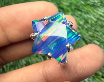Puede incluir: Anillo de plata con una gran ópalo en forma de estrella. El ópalo muestra iridiscencia azul, verde y naranja. El anillo se sostiene sobre un fondo verde borroso.