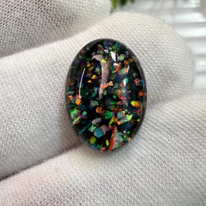May include: An oval-shaped cabochon gemstone featuring a deep black base embedded with a dense array of iridescent, multi-colored flecks in shades of red, green, blue, and orange, creating a vibrant, reflective, and highly textured appearance.