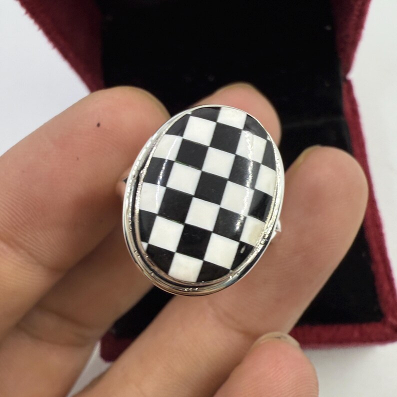 May include: Oval-shaped ring with a black and white checkered design. The ring has a silver-colored band and a large, eye-catching checkerboard pattern. The ring is held in a red velvet jewelry box.