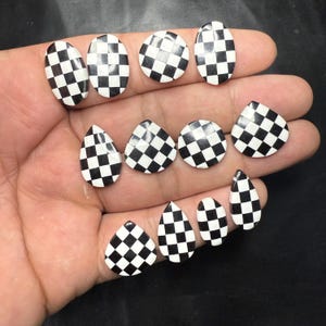 May include: A collection of black and white checkered nail art pieces in various shapes, including oval, round, and teardrop. The pieces are arranged on a hand, showcasing the contrasting pattern.