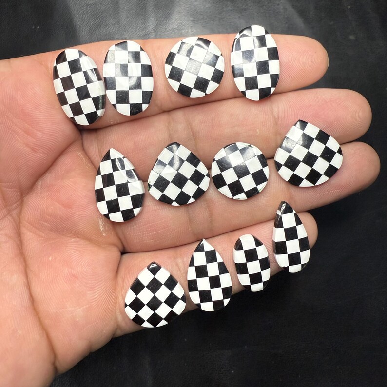 May include: A collection of black and white checkered nail art pieces in various shapes, including oval, round, and teardrop. The pieces are arranged on a hand, showcasing the contrasting pattern.