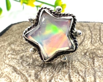Puede incluir: Un anillo en forma de estrella con una gran piedra preciosa iridiscente. La piedra preciosa muestra un arcoíris de colores. El anillo tiene una banda plateada texturizada con pequeños adornos redondos. El anillo está sobre una superficie texturizada de tono neutro.