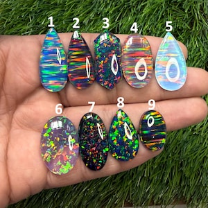 May include: A collection of nine polished opal gemstones in various shapes and colors. The opals display vibrant internal patterns of blue, green, orange, and purple. The gemstones are displayed on a hand against a green background.