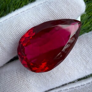 May include: A pear-shaped, faceted gemstone with a deep, vibrant red color. The stone features numerous triangular and diamond-shaped facets that reflect light, creating a brilliant, sparkling appearance across its polished, translucent surface.