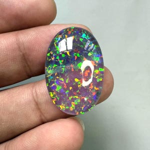 May include: An oval-shaped black opal gemstone with vibrant flashes of green, blue, and orange. The gemstone is held in a hand, showcasing its polished surface and internal play of color. The opal's dark base enhances the brilliance of the colorful inclusions.