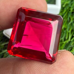 Puede incluir: Una piedra preciosa de color rojo vibrante con corte cuadrado y superficie facetada que refleja la luz. La piedra presenta bordes afilados y definidos, con un tono carmesí profundo y saturado en todo su cuerpo transparente y geométrico.