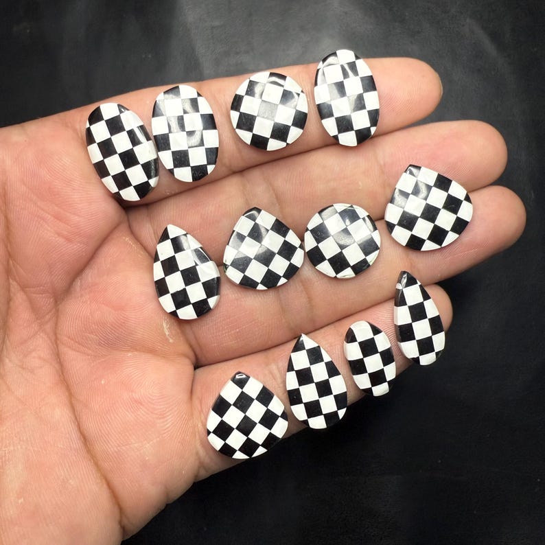 May include: A collection of black and white checkered nail art pieces in various shapes, including oval, round, teardrop, and triangular designs. The pieces are displayed against a dark background.