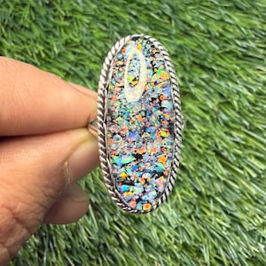 Op de afbeelding: Een ovale ring met een zilveren band en een kleurrijke, mozaïekachtige steen. De steen heeft een mix van blauwe, groene, oranje en zwarte fragmenten, omlijst door een zilveren, touwachtige rand. De ring wordt tegen een groene achtergrond getoond.