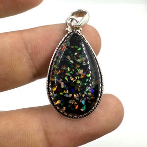 Puede incluir: Un colgante en forma de lágrima con una piedra de ópalo negro. La piedra está llena de motas iridiscentes de color, incluyendo verde, azul y naranja. El colgante tiene un engaste de metal plateado y un lazo en la parte superior para una cadena.