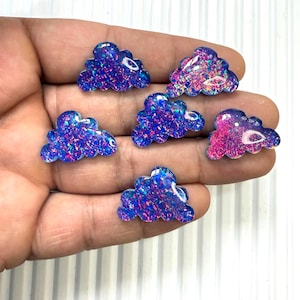 May include: Seven cloud-shaped resin embellishments with iridescent glitter. The clouds are primarily blue with hints of pink and purple. These decorative pieces are ideal for crafting and jewelry making.